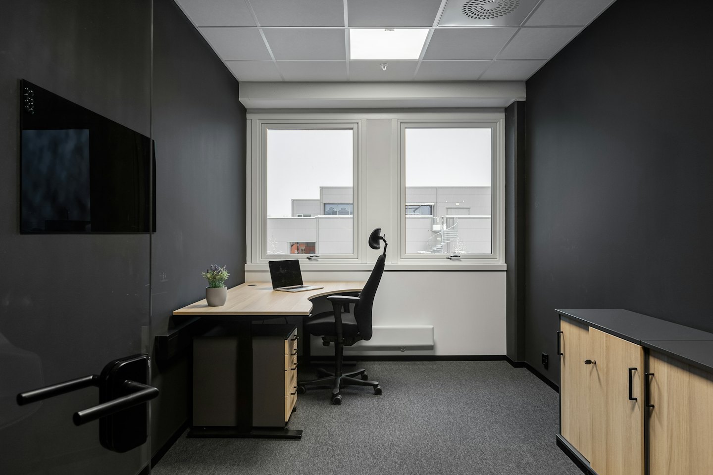 Modern office with a wooden desk and laptop near windows.
